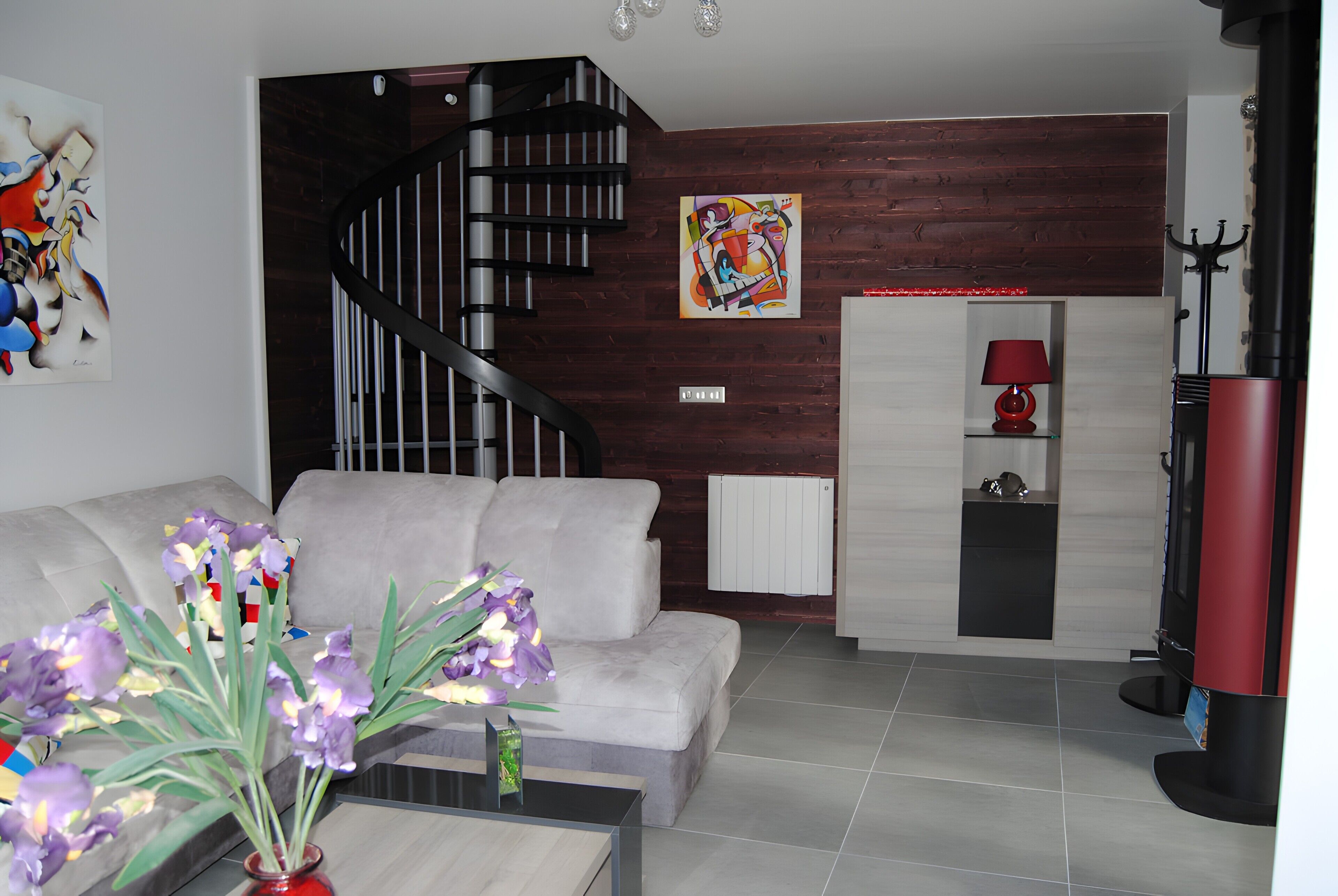 A modern living room with a grey sofa, a red vase with flowers, a black spiral staircase, a colorful abstract painting, and a red lamp on a white cabinet.