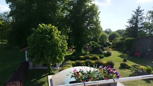 A balcony with a metal railing, a flower pot with flowers, and a view of a well-maintained garden with various plants and a wooden structure in the background. - Gîte 'Des Taravelles' with Private Pool, Private Terrace and Wi-Fi (Manzat)