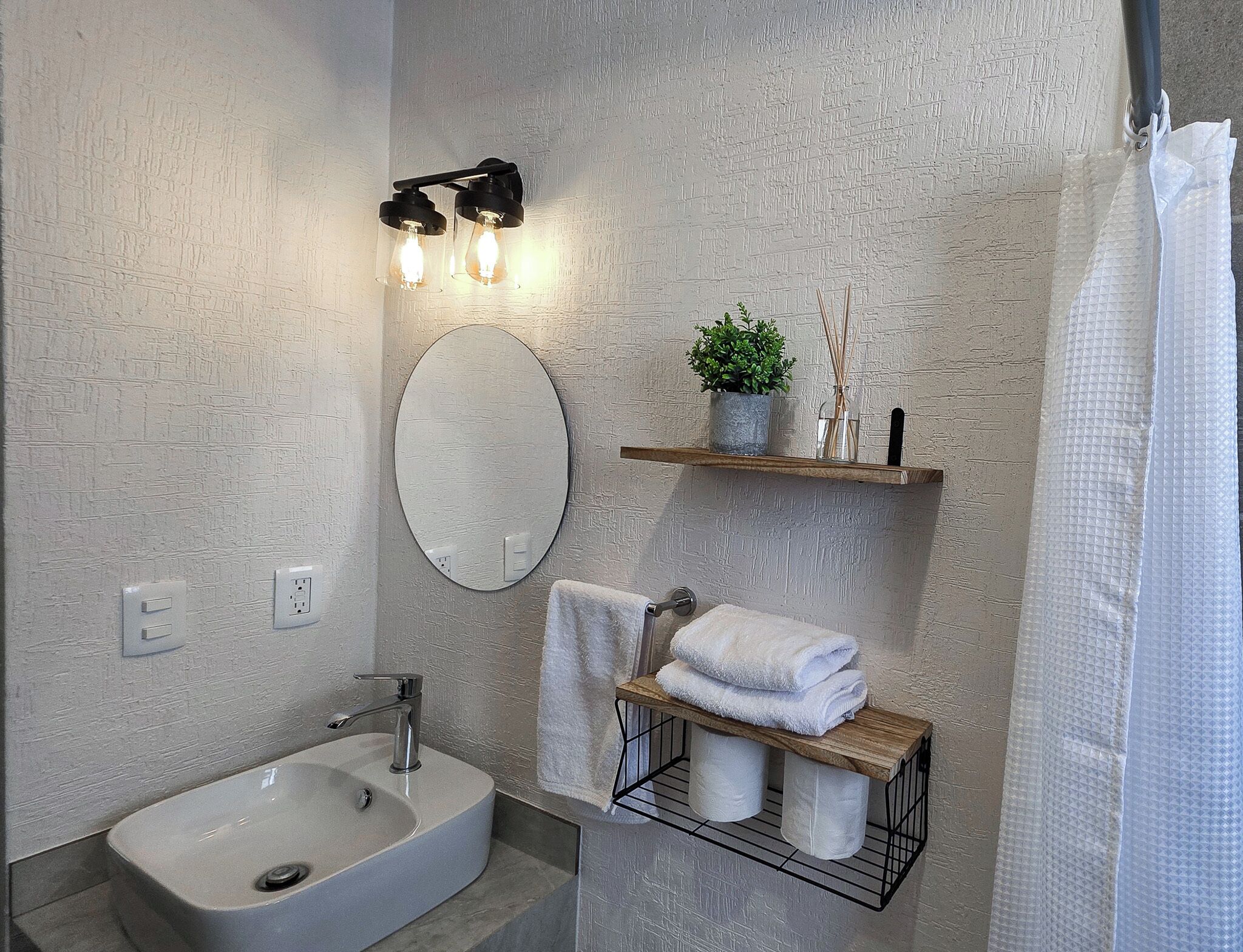 A modern bathroom with a white sink, a mirror, a towel rack, and a shelf with toiletries.