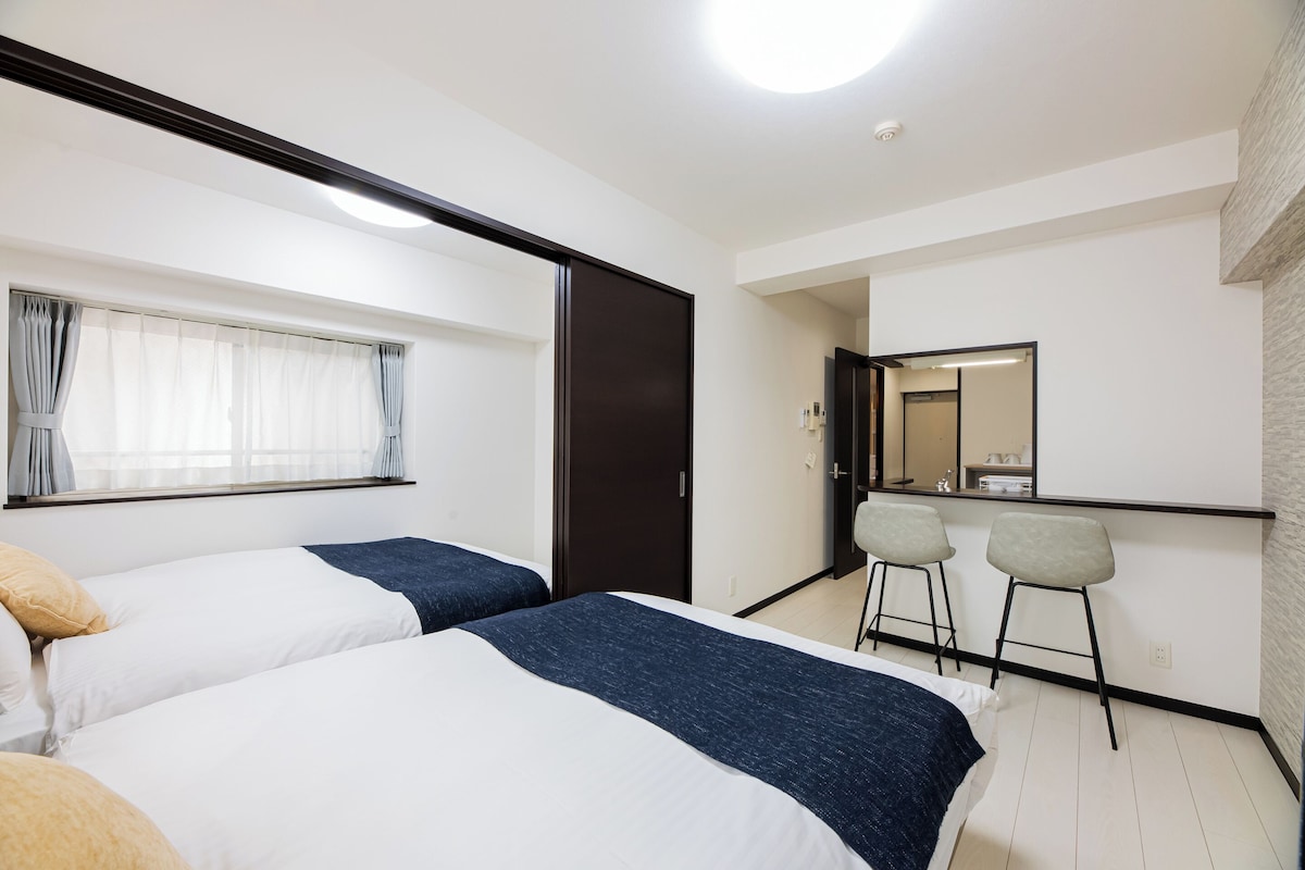A hotel room with two beds, a small desk with two chairs, a mirror, and a window with curtains.