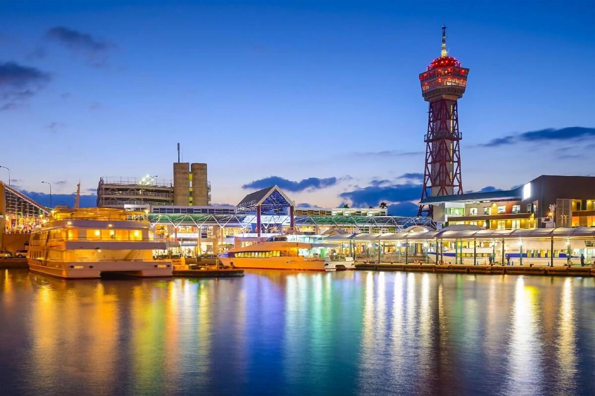 Un paisaje urbano frente al mar con una torre destacada, un gran crucero atracado y edificios modernos con fachadas iluminadas.