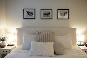 A neatly made bed with white linens and multiple pillows, a bedside table with a lamp and a small plant, and three framed pictures on the wall above the bed.