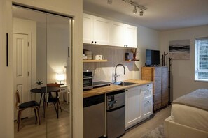 A compact kitchen with white cabinets, a dishwasher, a microwave, and a sink. There's a dining area with a table and chairs, and a bed in the corner.