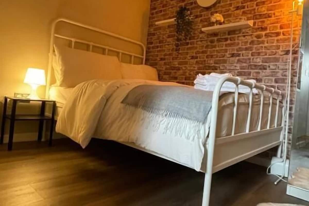 Una cama individual con estructura metálica, una mesita de noche con lámpara, una pared de ladrillo y un pequeño estante con una planta.
