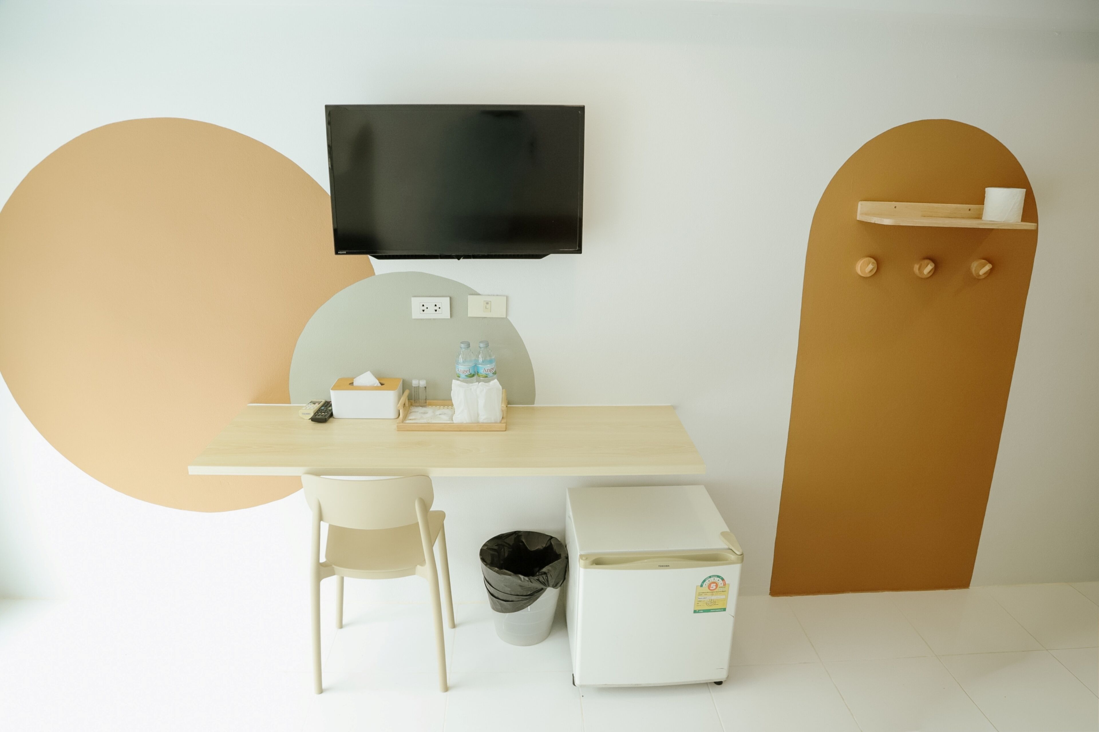 Een moderne kamer met een flatscreen televisie aan de muur, een klein tafeltje met een stoel, een prullenbak en een compacte koelkast.