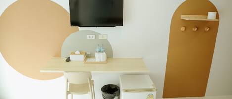 Een moderne kamer met een flatscreen televisie aan de muur, een klein tafeltje met een stoel, een prullenbak en een compacte koelkast.