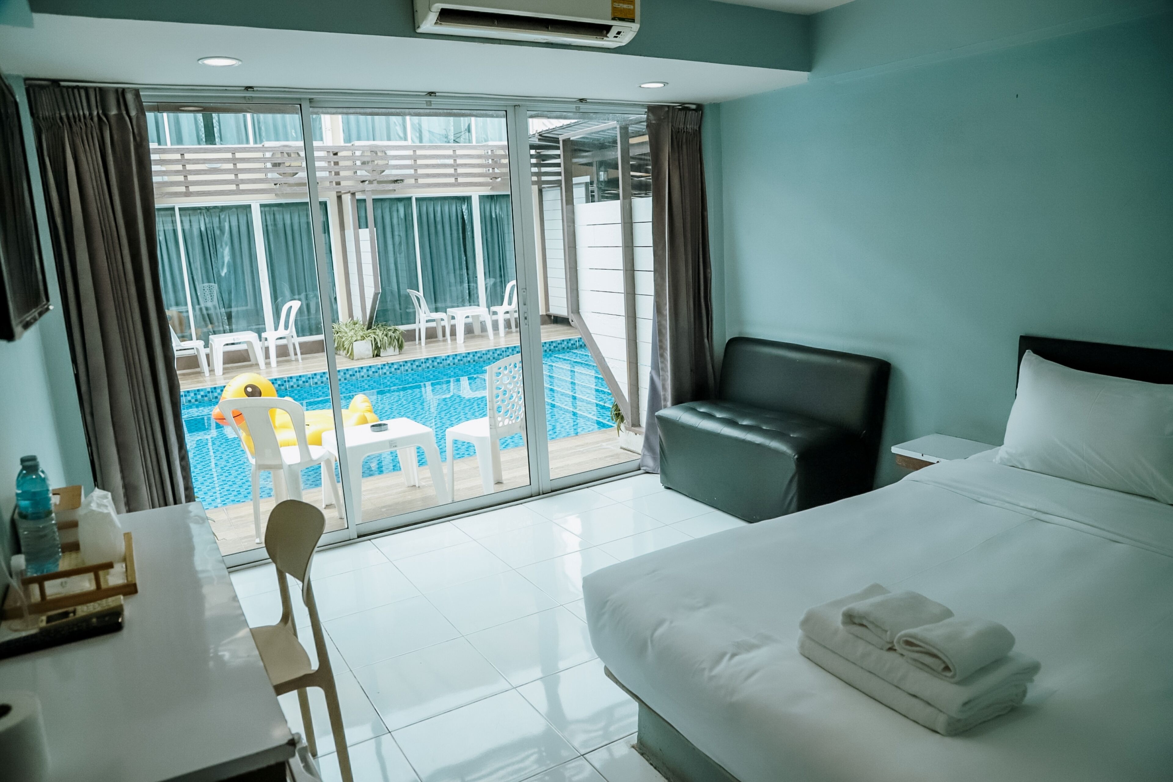 A hotel room with a bed, a desk, a chair, a small table with a bottle and glasses, a nightstand, and a view of a pool and outdoor seating.