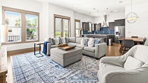 A spacious living area with a grey sofa, a coffee table, and a patterned rug. There is a kitchen in the background with modern appliances and a dining area.