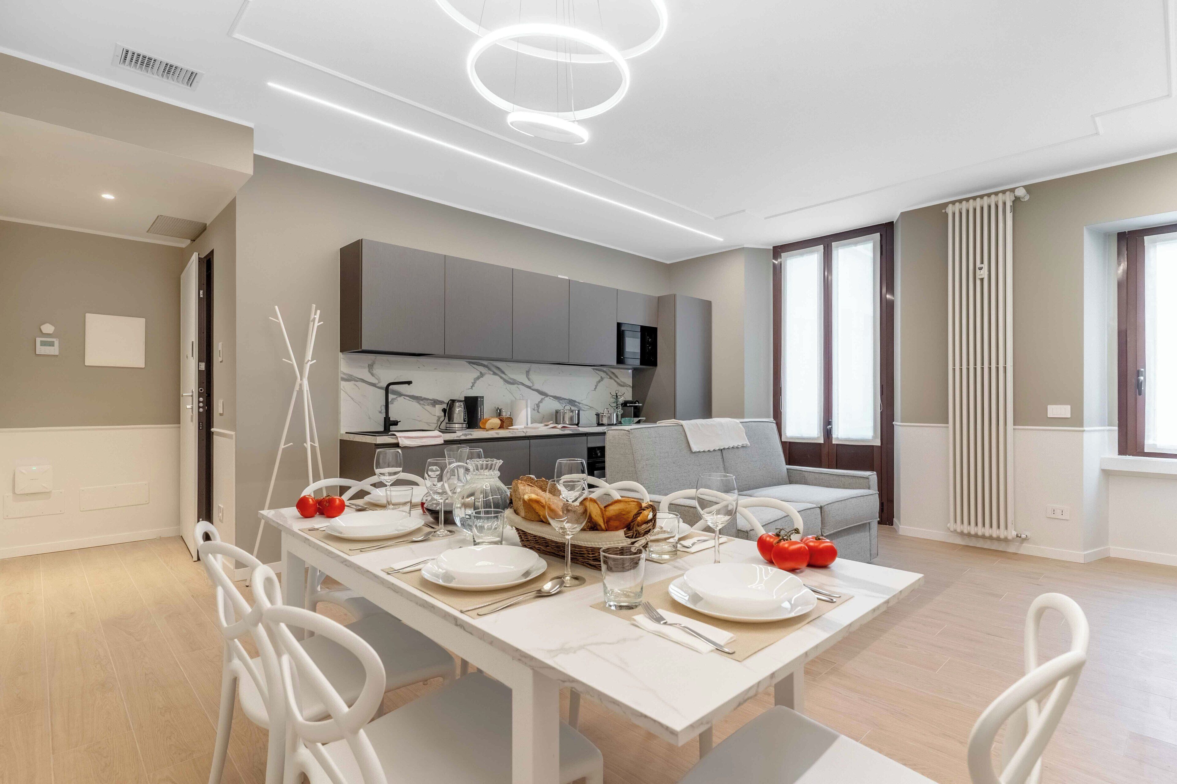 Una cocina moderna con electrodomésticos integrados, un comedor con una mesa blanca dispuesta para seis personas, y un sofá gris al fondo.