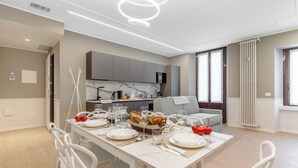 A modern kitchen with built-in appliances, a dining area with a white table set for six, and a grey sofa in the background. - Appartamento Puccini - La Vista Apartments (Milan)