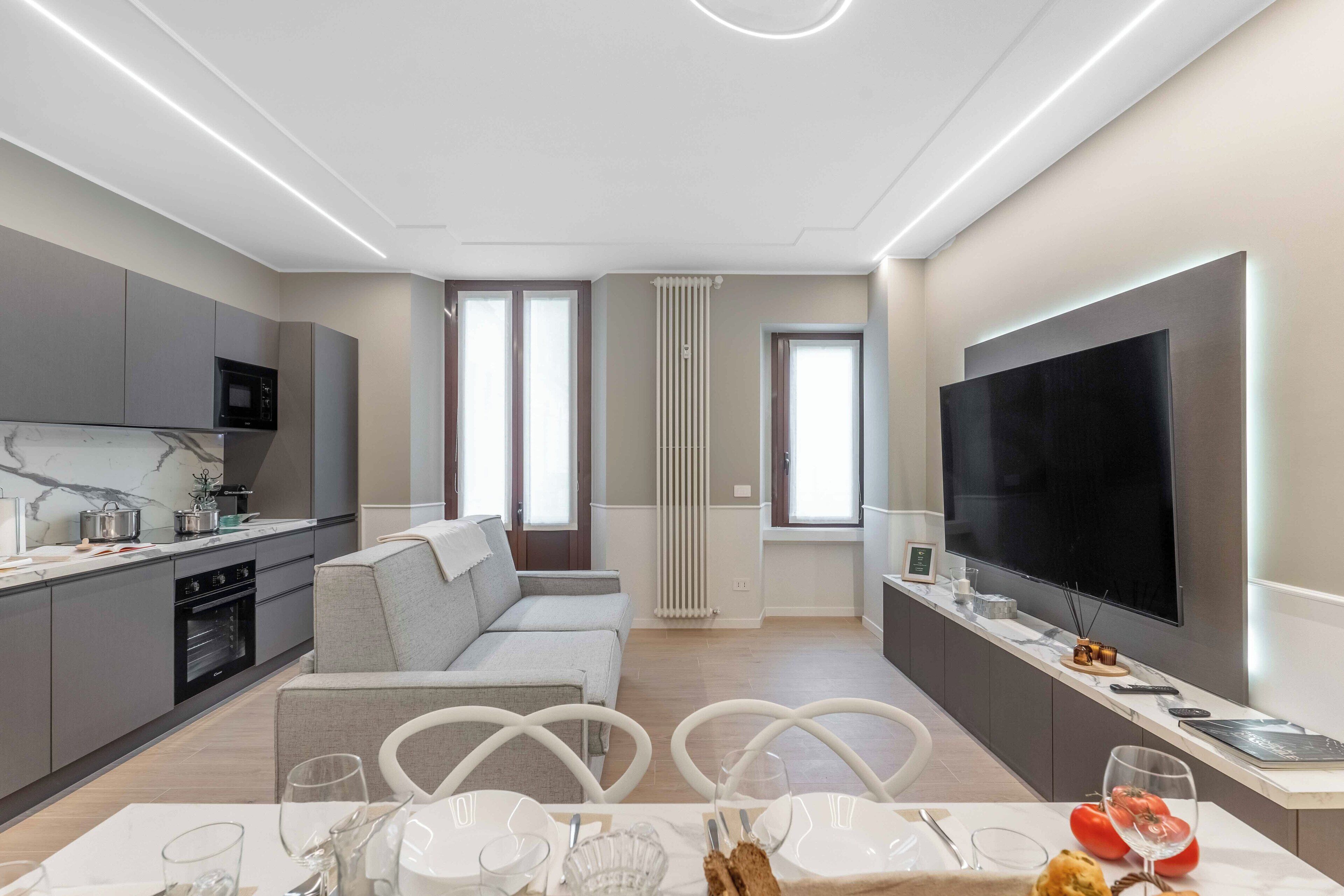 Une cuisine moderne dotée d’un four et d’un micro-ondes encastrés, un coin repas avec une table préparée pour deux personnes, et un grand téléviseur à écran plat fixé au mur.