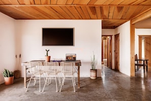 A room with a wooden ceiling, a flat-screen TV mounted on the wall, a dining table with chairs, and potted plants.