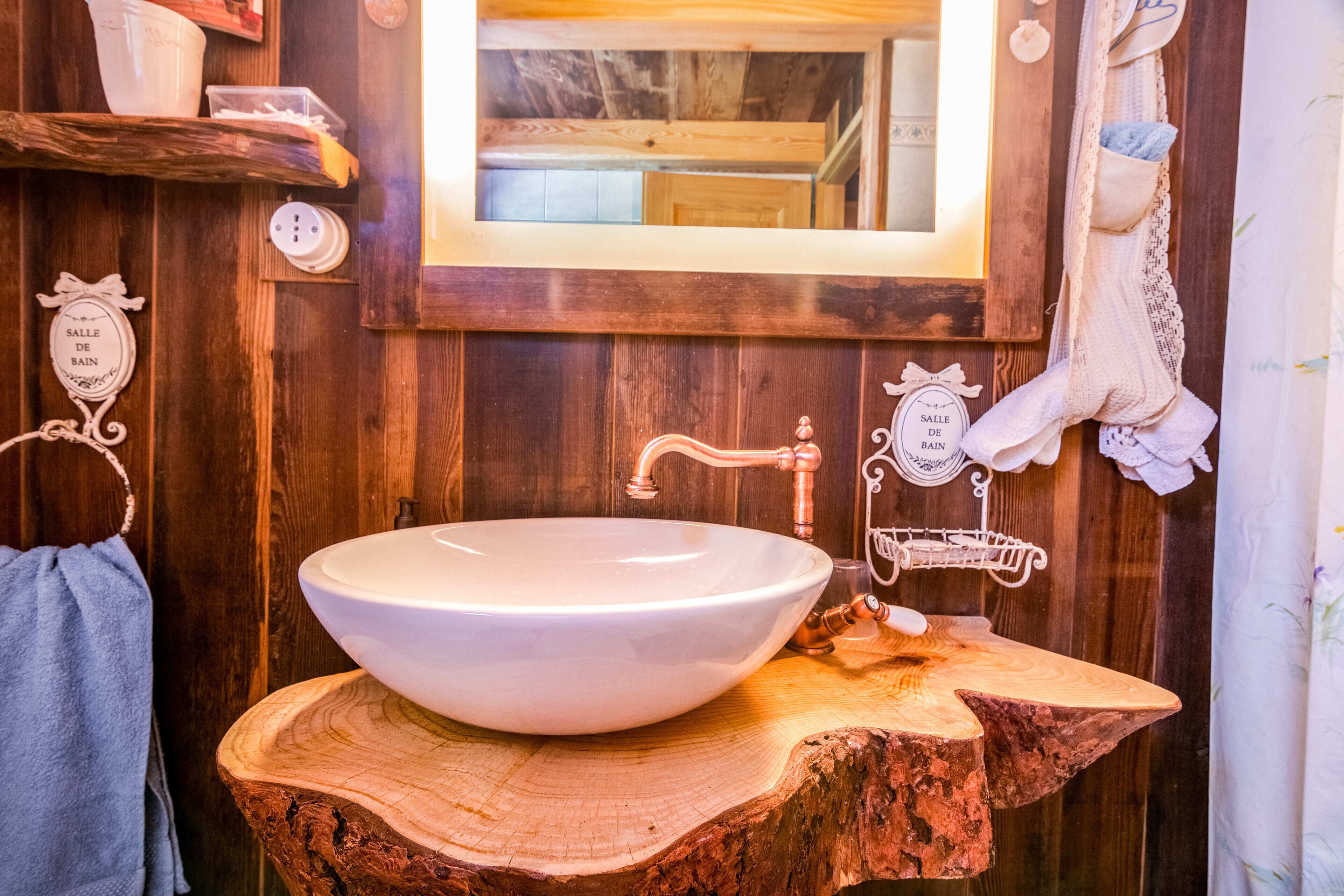 Ein rustikales Badezimmer mit einem großen, weißen Waschbecken auf einem hölzernen Baumstumpf, Holzwänden und einem Spiegel über dem Waschbecken.