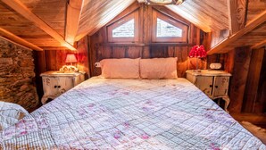 Une chambre avec un plafond en bois, un mur en pierre, un lit recouvert d’une courtepointe à motifs floraux et une table de chevet avec une lampe.