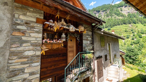 Un bâtiment rustique en pierre, doté d’une porte d’entrée en bois, d’un balcon avec une rambarde en métal et offrant une vue sur une vallée verdoyante.