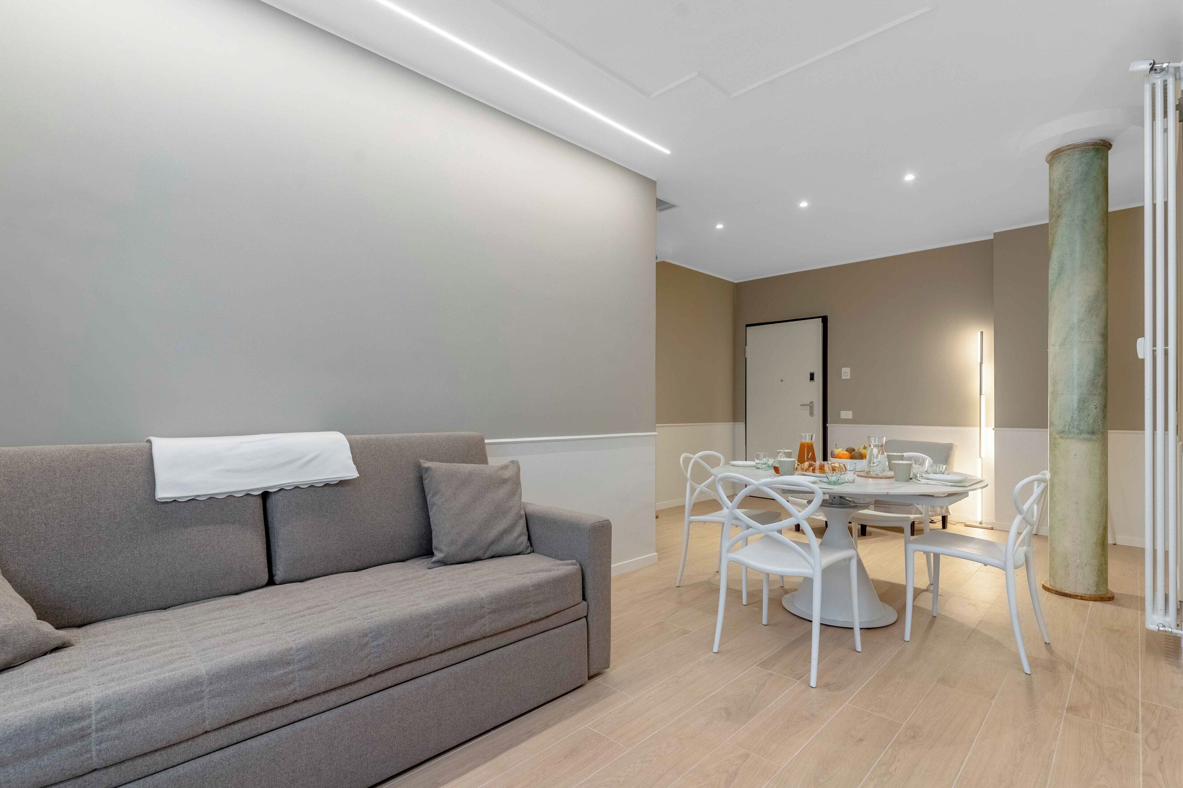 A modern living room with a grey sofa, white chairs, a round dining table, and a large mirror.