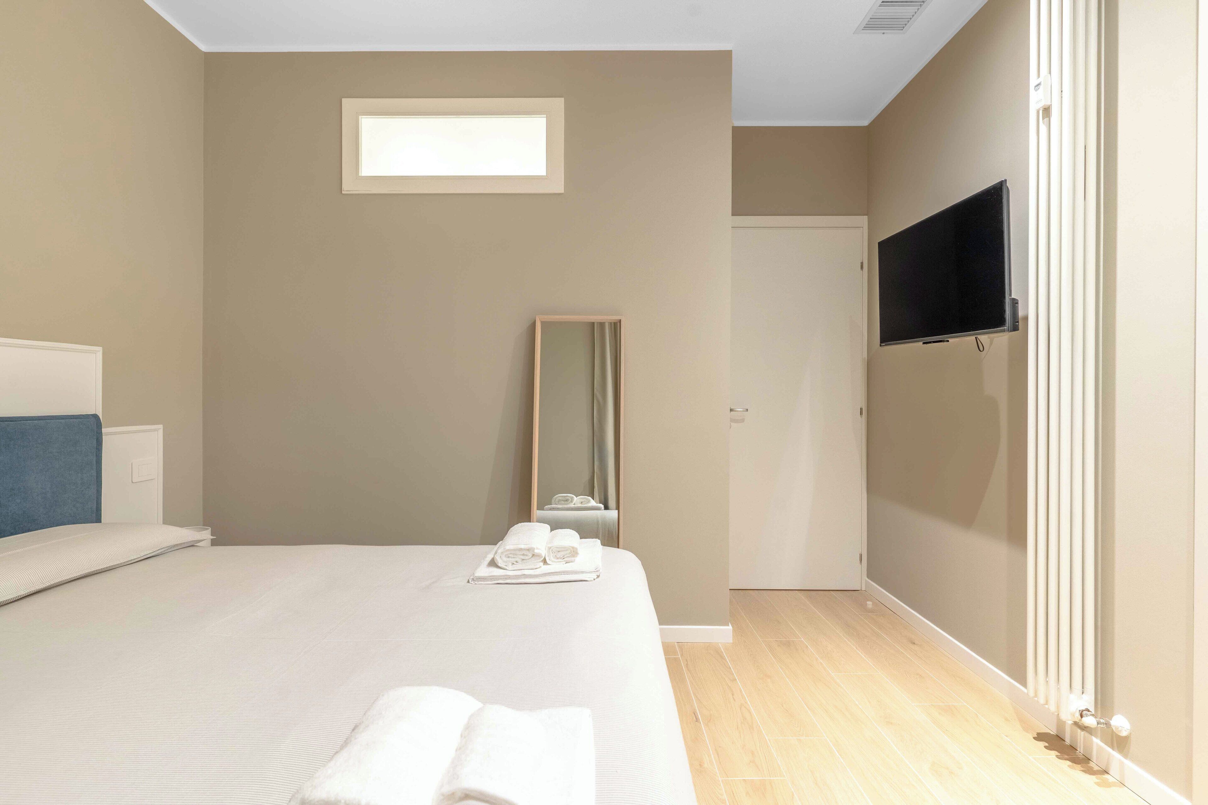 A bedroom with a bed, a flat-screen TV, a mirror, and a window.