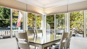 Een eethoek met een grote tafel en zes stoelen, grote ramen met een Amerikaanse vlag, en uitzicht op een parkeerplaats en bomen.