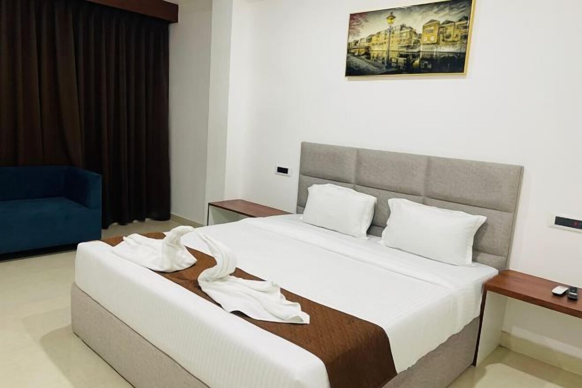 A neatly made bed with white linens and a brown accent, a blue armchair, and a painting on the wall.