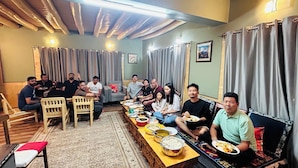 Un grupo de personas sentadas alrededor de una mesa larga con diversos platos, en una habitación con vigas de madera en el techo y cortinas grises.