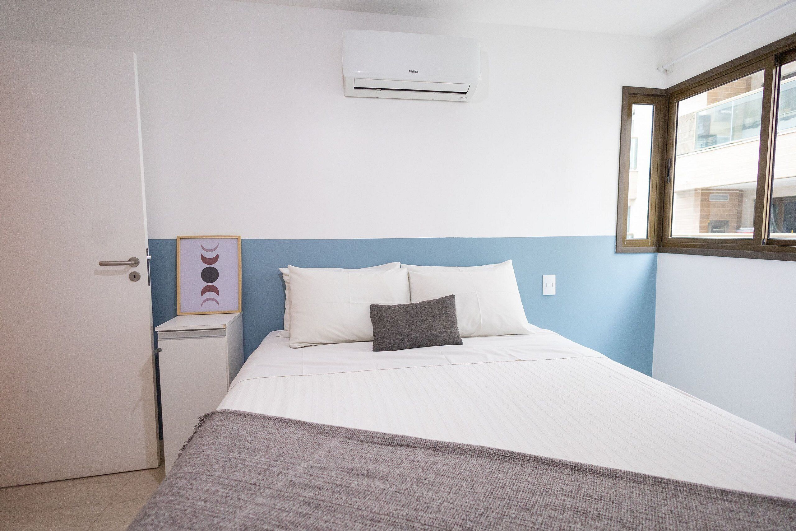 Een slaapkamer met een bed, nachtkastje, airconditioning, een ingelijste foto en een raam.