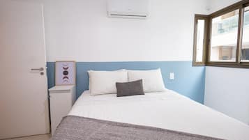 Une chambre à coucher comprenant un lit, une table de chevet, un appareil de climatisation, un tableau encadré et une fenêtre.