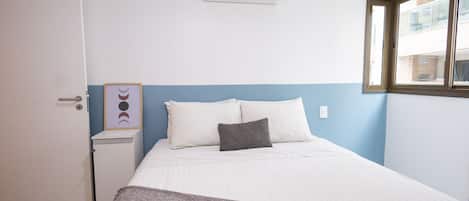 A bedroom with a bed, a nightstand, an air conditioning unit, a framed picture, and a window.