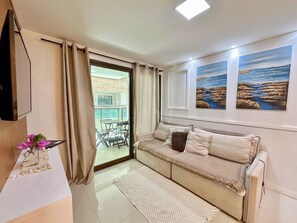 A living room with a sofa, a TV, a window with curtains, and artwork on the wall. - Habitat Hospedagens (Arraial do Cabo)