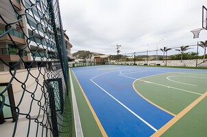 Quadra de basquete ao ar livre com cercas de arame, cesta de basquete e um edifício ao fundo.