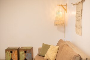 Quarto com sofá bege, travesseiro verde e estampado, um pendente de macramé na parede e dois banquinhos de madeira com rostos pintados.