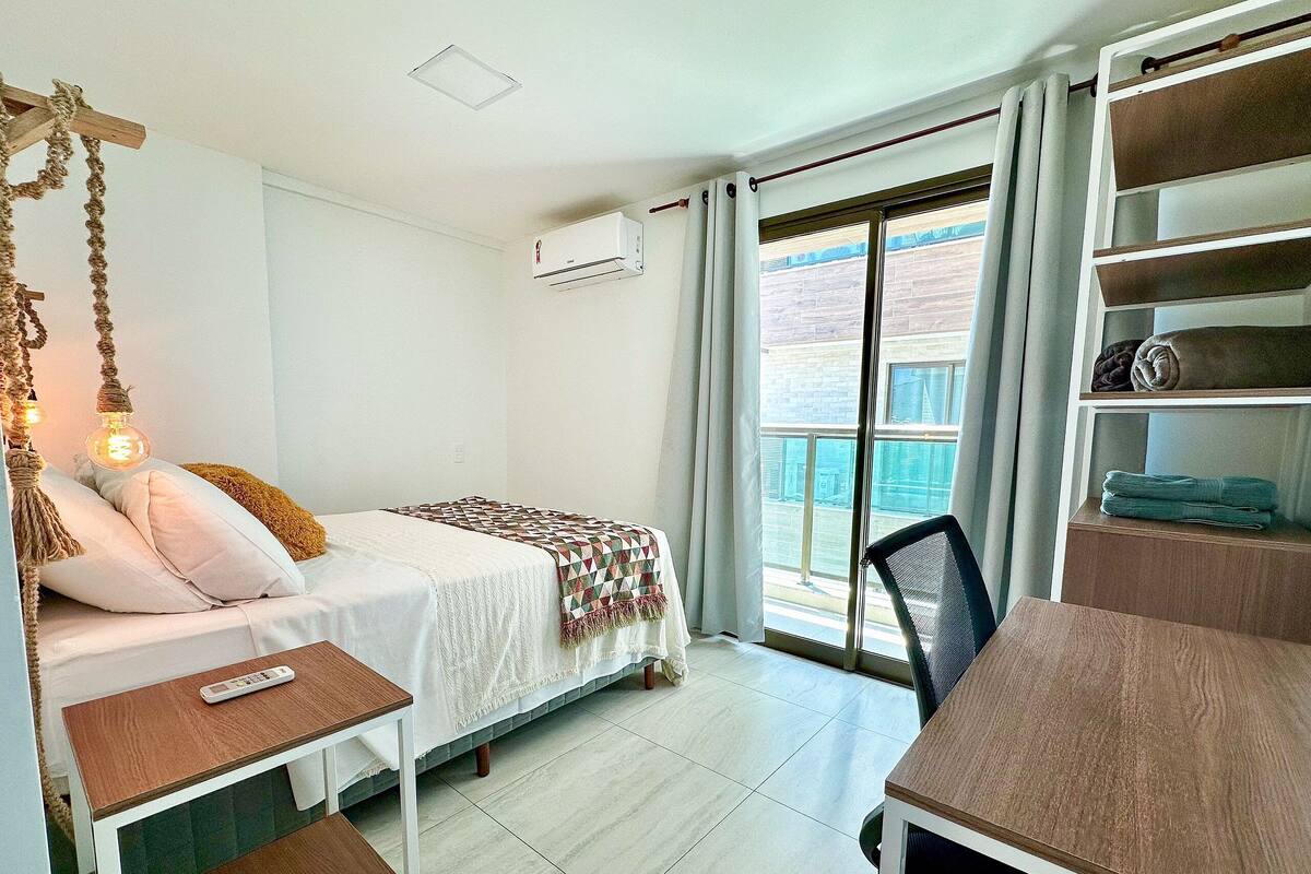 A room with a bed, a desk, a chair, a hanging light fixture, a window with curtains, and a wall-mounted air conditioner.