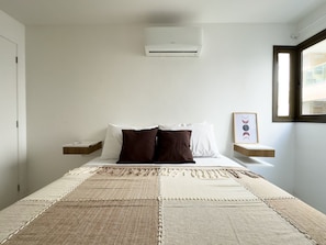 A bedroom with a bed, pillows, a bedside table, a wall-mounted air conditioner, a window, and a framed picture. - Habitat Hospedagens (Arraial do Cabo)