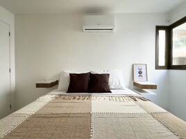 Quarto com cama, almofadas, mesa de cabeceira, ar-condicionado fixado na parede, janela e um quadro emoldurado.