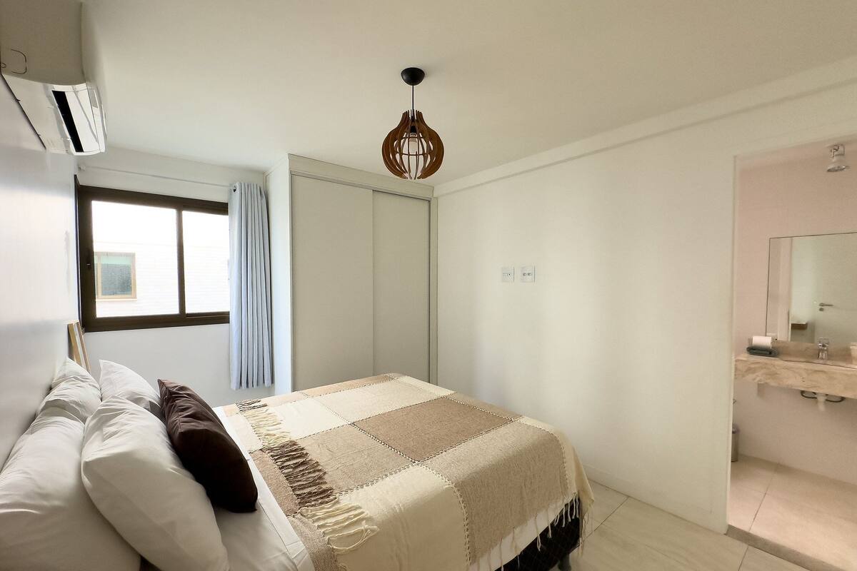 A bedroom with a bed, a window, a wardrobe, a hanging light fixture, and a bathroom with a mirror.