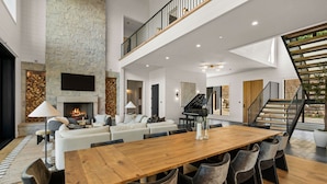 Un vaste espace de vie comprenant une grande table à manger en bois, des sièges confortables, une cheminée et un escalier menant à un niveau supérieur.
