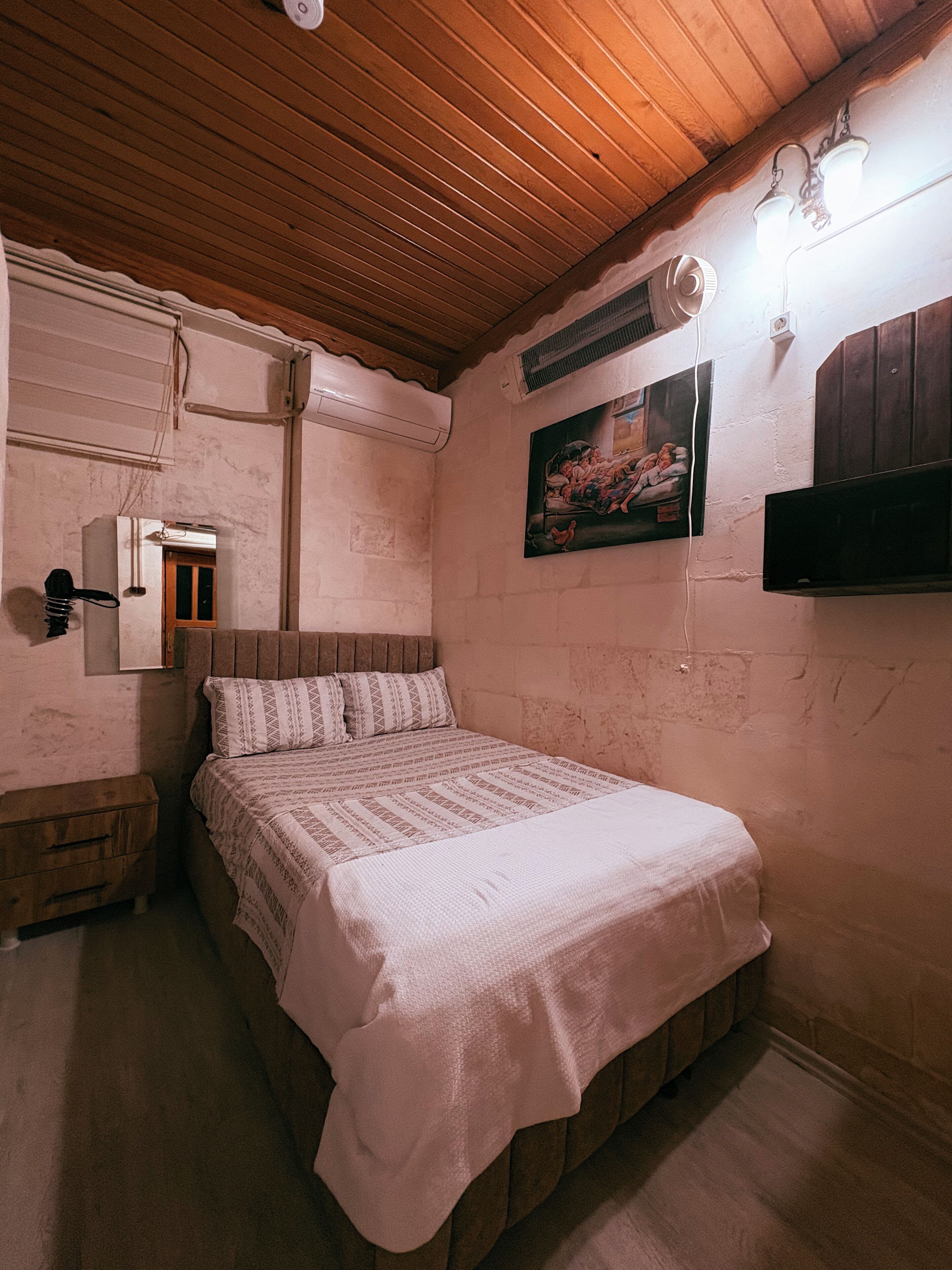 A bedroom with a wooden ceiling, a bed with a white bedspread, a wooden nightstand, a wall-mounted TV, and a painting.