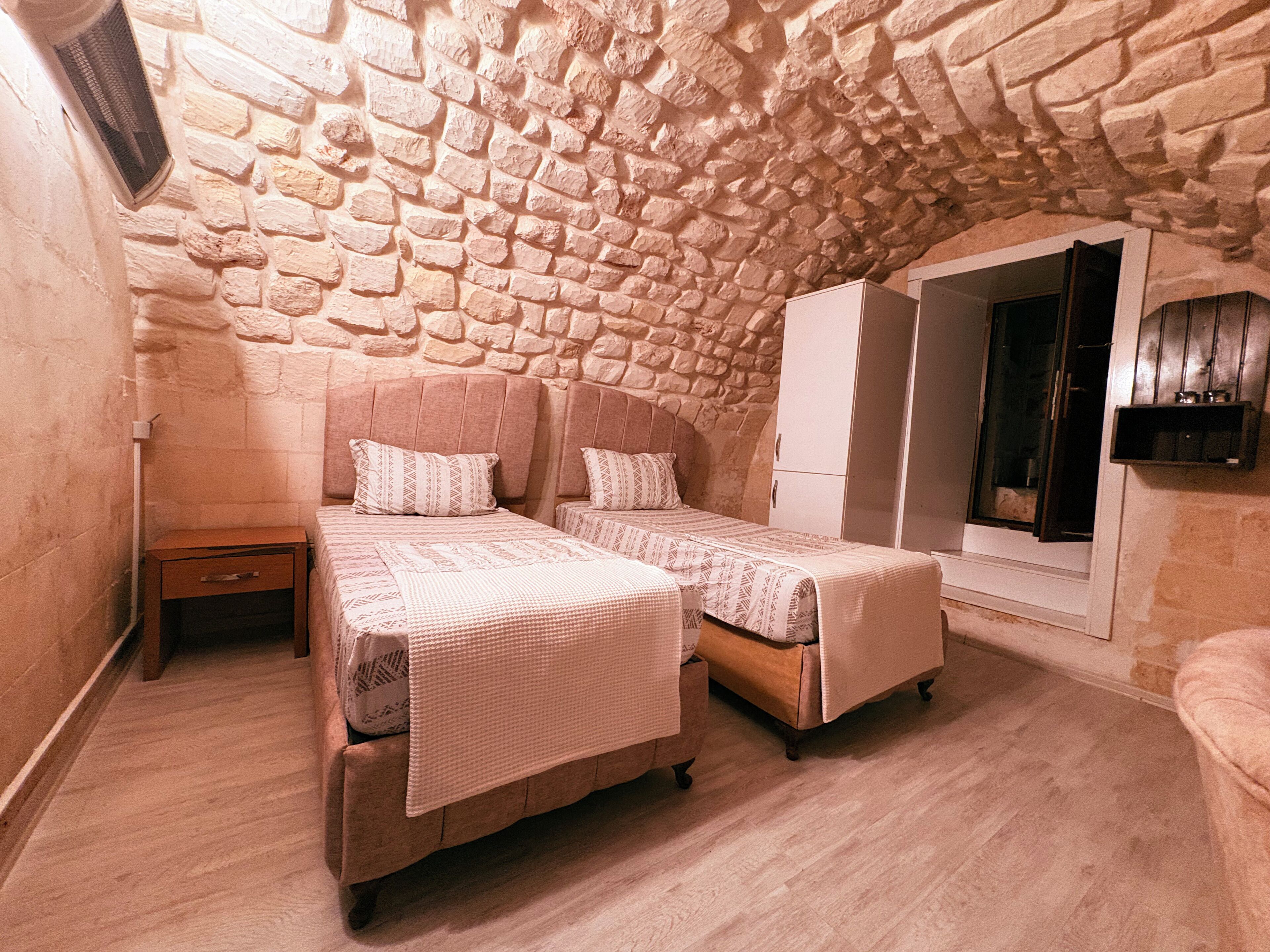 A bedroom with two beds, a wooden wardrobe, and a stone wall.