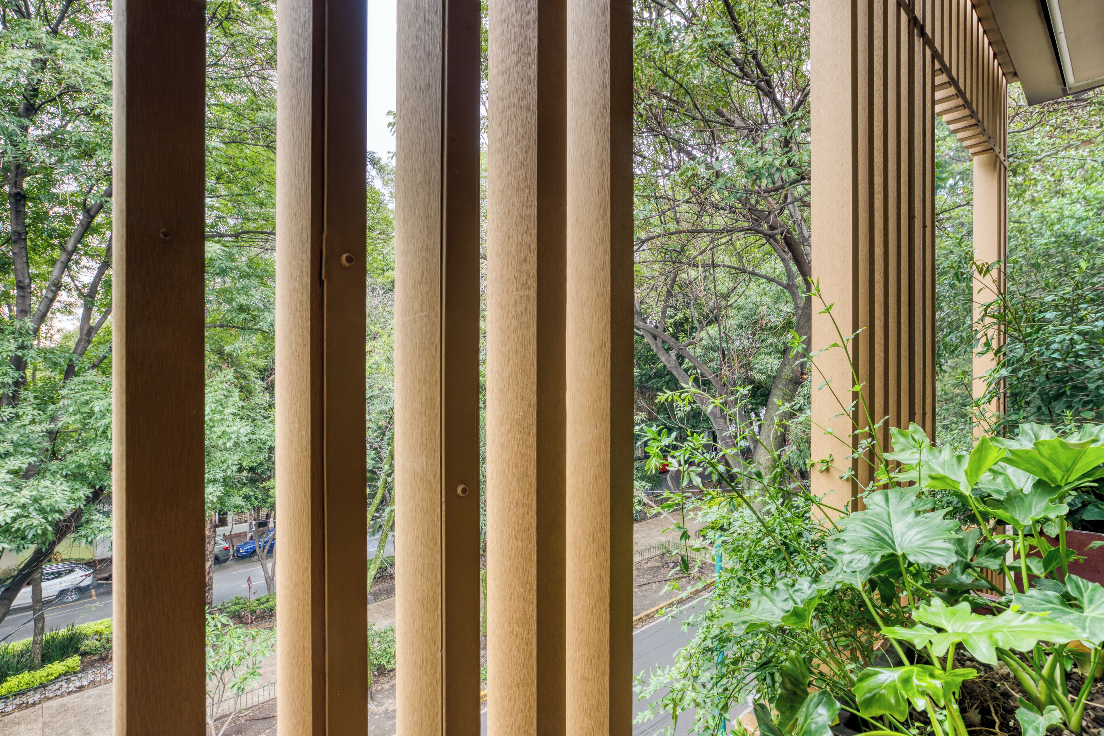 A view through wooden slats of a balcony with greenery and a road in the background.