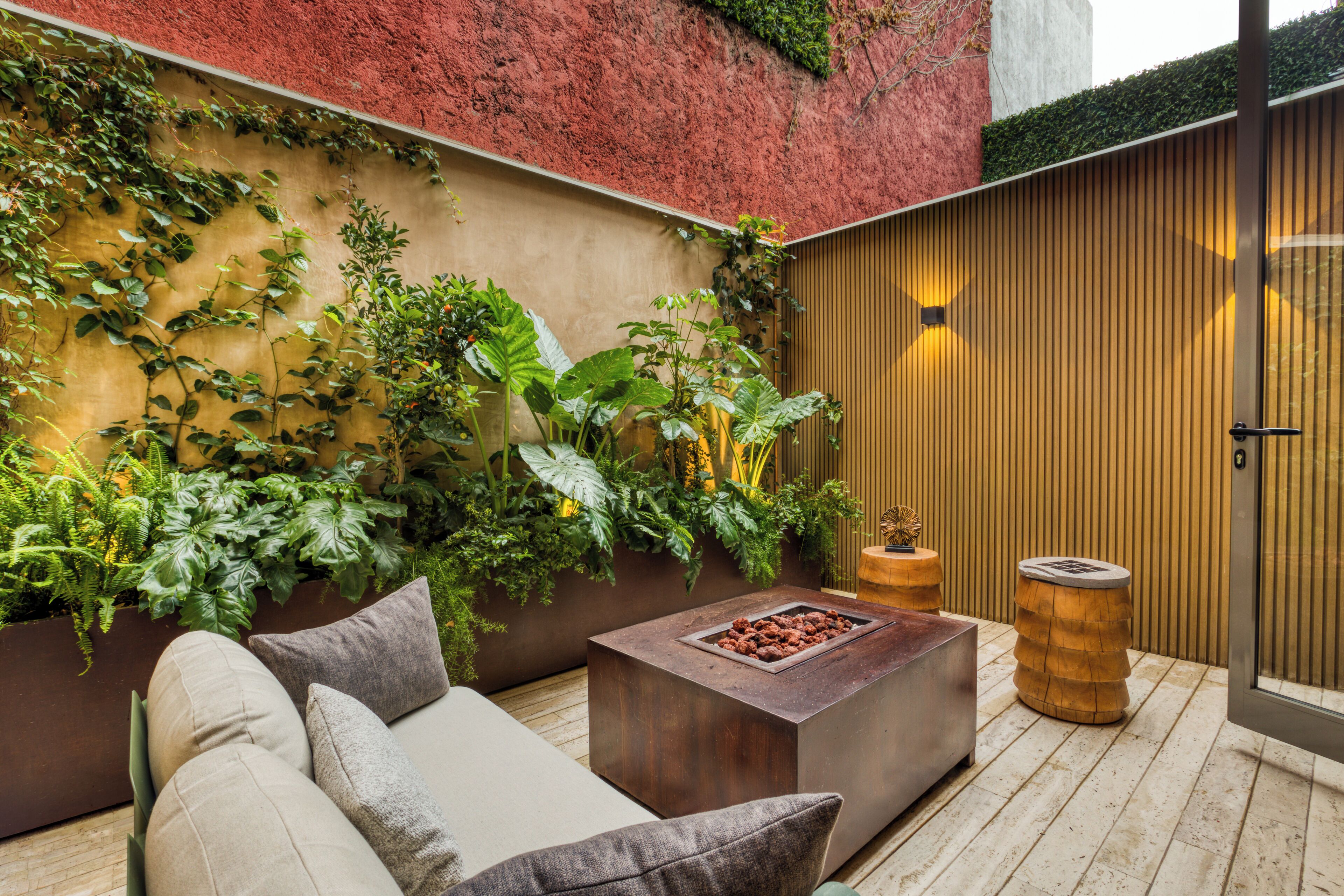 A cozy outdoor seating area with a cushioned sofa, a wooden coffee table, and a planter wall with various plants.