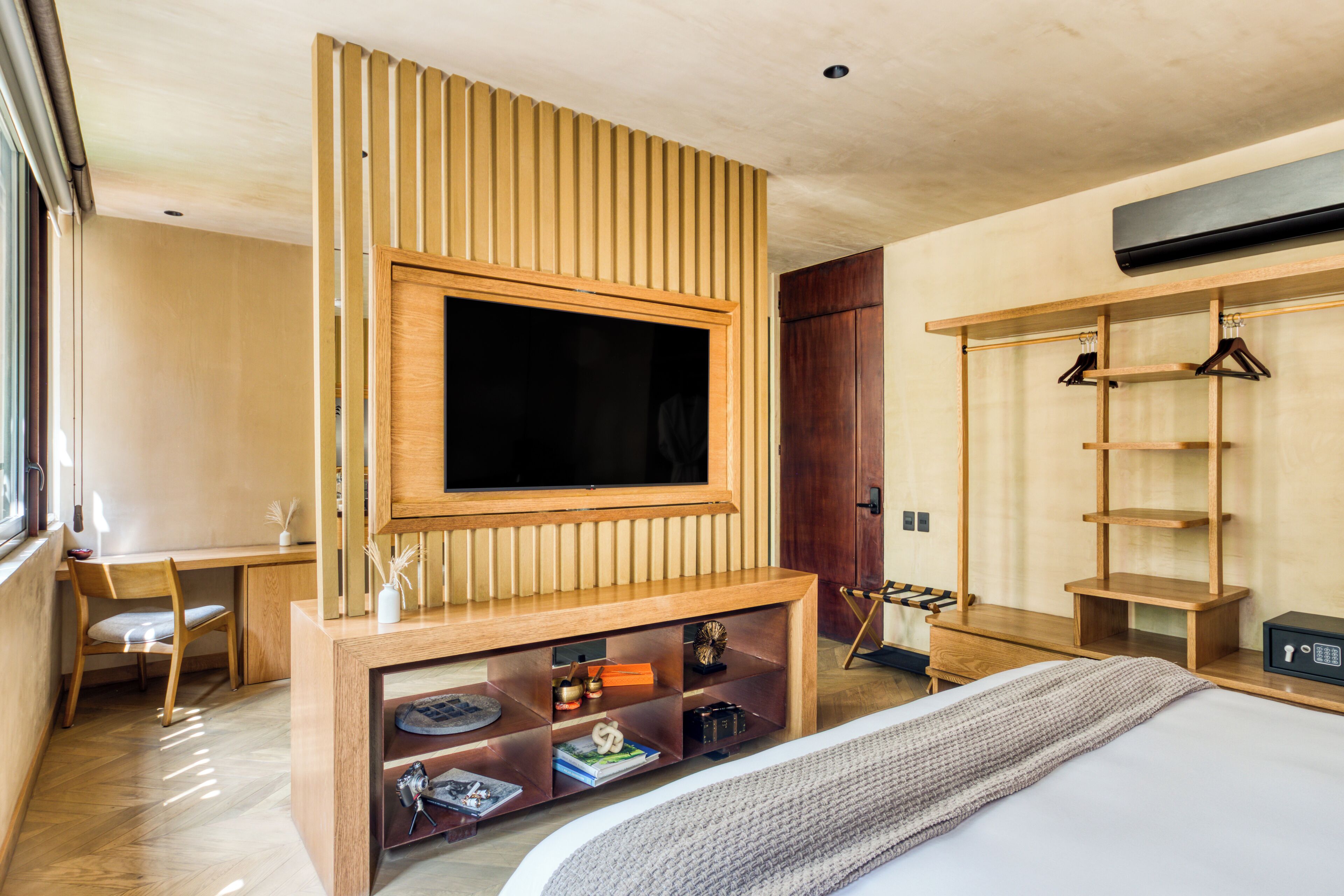 A modern hotel room with a large flat-screen TV mounted on a wooden panel, a wooden wardrobe, a small desk, and a bed with a knitted throw.