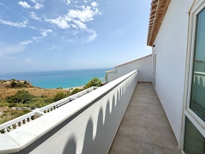 A balcony with a view of the sea, a white building, and a tiled roof.