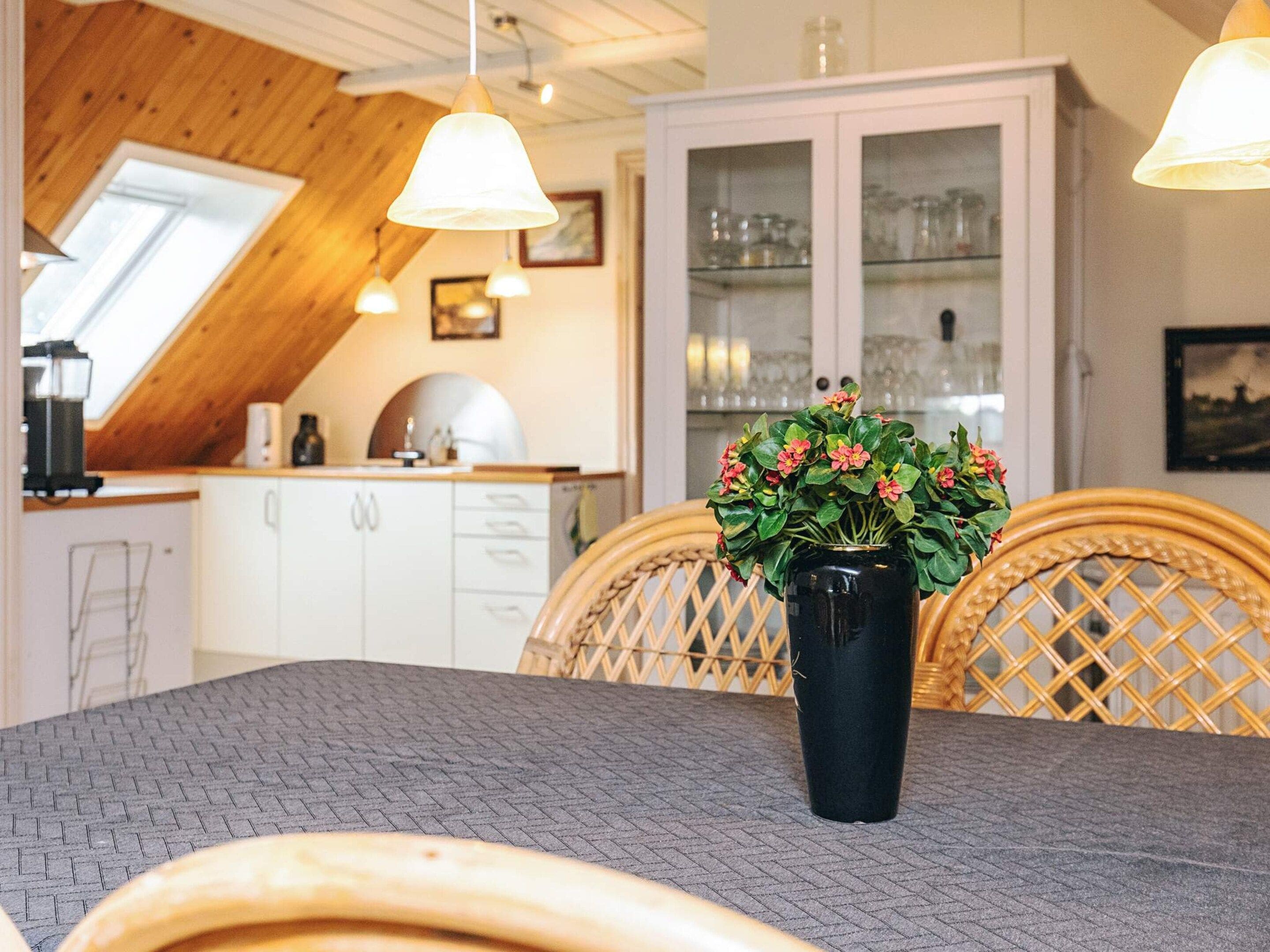 A dining area with a table, chairs, a vase with flowers, and a kitchen in the background.