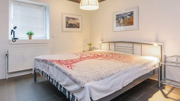 Een slaapkamer met een groot bed, een radiator, een raam met zonwering, een hanglamp en twee ingelijste foto's aan de muur.