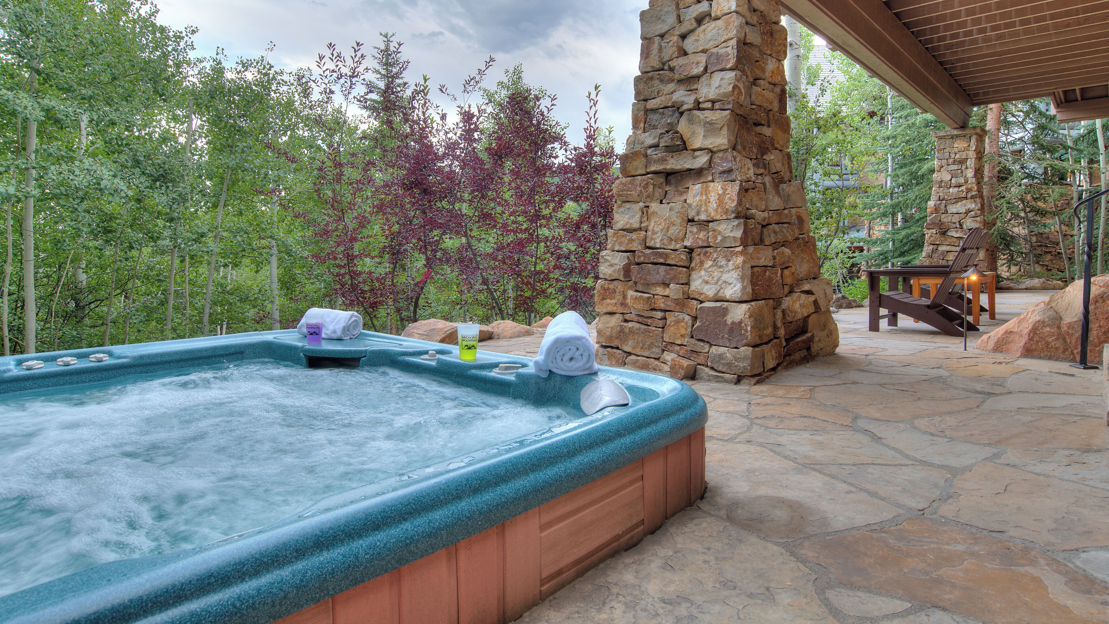 A hot tub with a stone pillar and wooden deck, surrounded by trees and a stone patio.