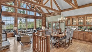 A spacious living area with a high ceiling and large windows, a dining area with wooden furniture, and a kitchen with wooden cabinets and a large island. - Luxury Ski-in/out Retreat | Timber Haven (Avon)