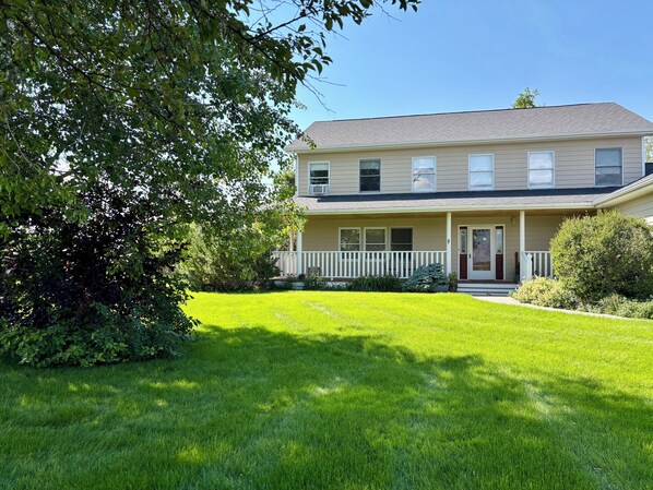 A two-story house with a front porch, a well-maintained lawn, and surrounding greenery.