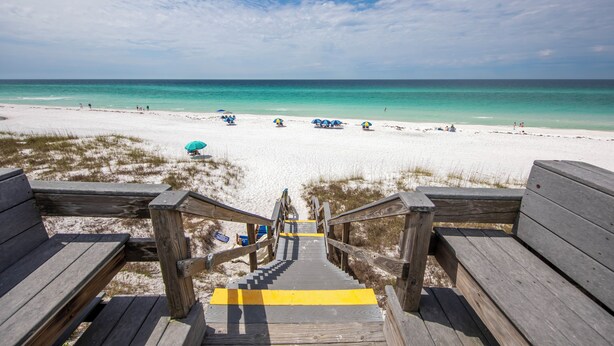 Eine Holztreppe führt zu einem Strand mit weißem Sand und klarem, türkisblauem Wasser.