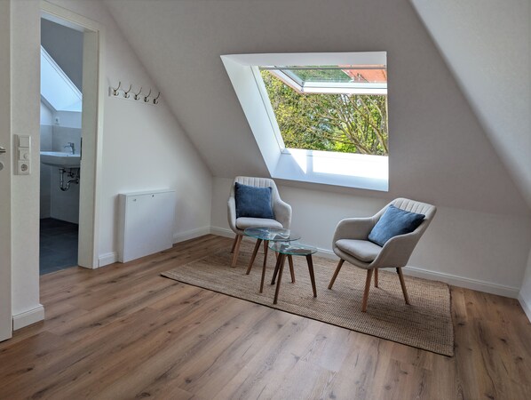 A room with a sloped ceiling, a skylight, two armchairs, a glass coffee table, and a view of greenery outside.