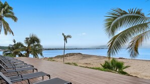 Una terraza de madera con sillones de descanso con vistas a una playa con palmeras y un cielo azul despejado.
