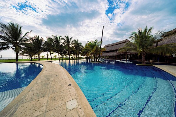 A large outdoor swimming pool with palm trees and a thatched-roof structure.